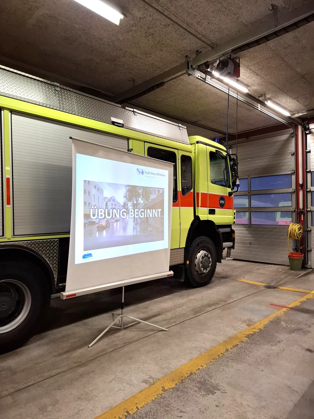 Überblick verschafft, Einsatzkräfte bereit, Übung beginnt. Eine Leinwand steht vor einem Feuerwehrauto.